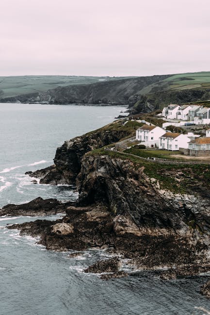 Drone view of small settlement located on rocky coast near rippling sea and hilly terrain against cloudless sky in nature